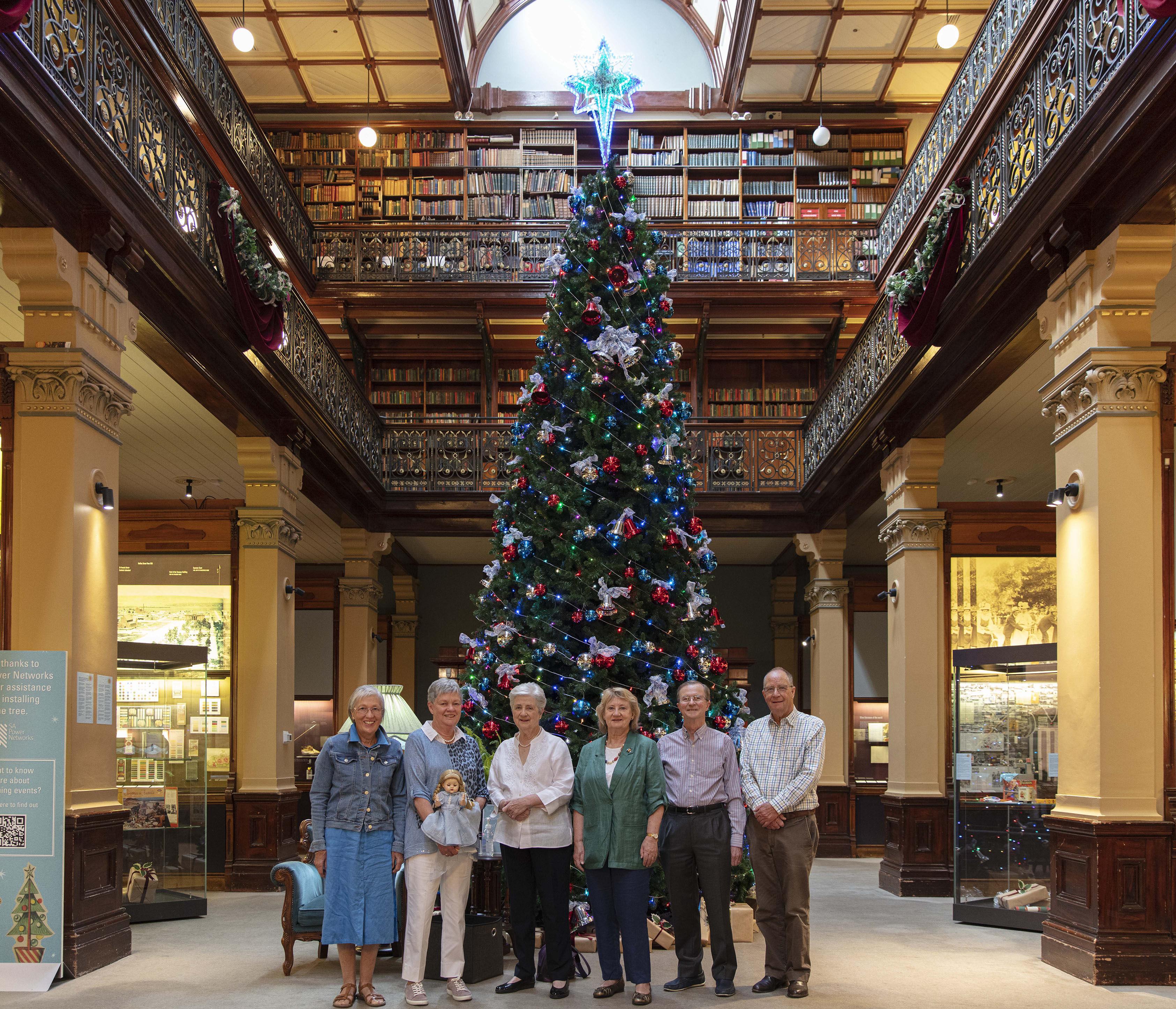 Six people standing in front of a large Christmas tree in the Mortlock Chamber.