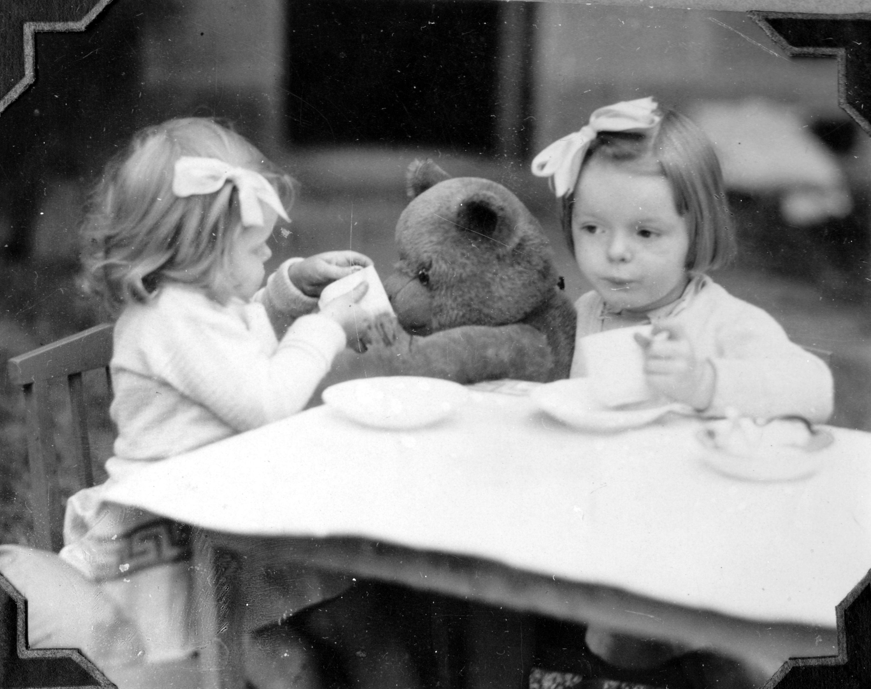 Two young children playing at a tea party. One child is offering tea to a teddy bear toy. 