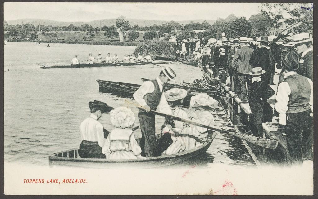 Crowd watching two rowing boats on lake, one with four ladies in large floral hats and man in straw boater pushing off from bank with oar.1915