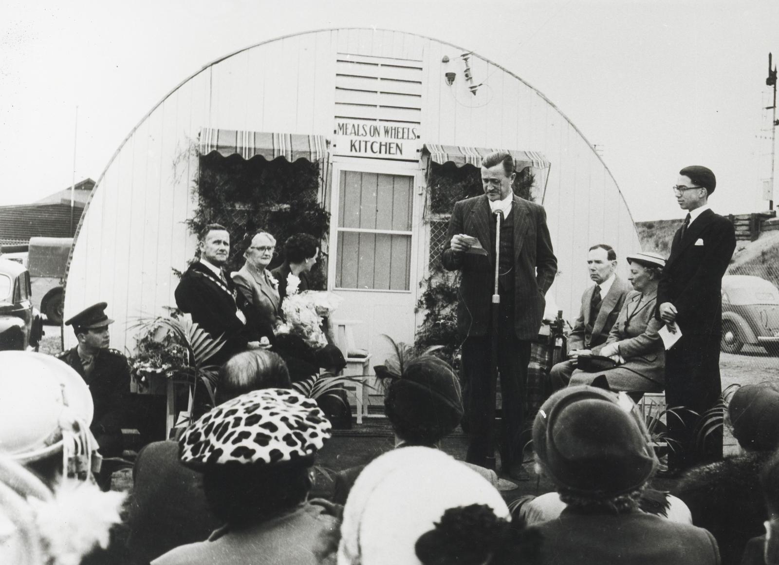 Audience watch speakers in front of Nissan Hut at opening of first Meals on Wheels kitchen, SA