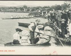 Crowd watching two rowing boats on lake, one with four ladies in large floral hats and man in straw boater pushing off from bank with oar.1915