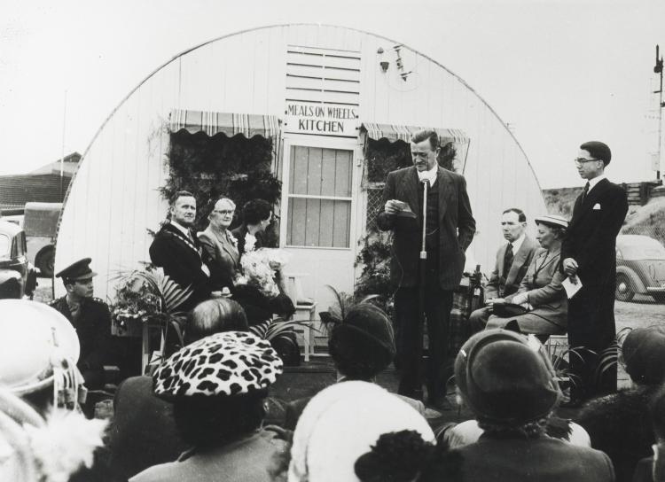 Audience watch speakers in front of Nissan Hut at opening of first Meals on Wheels kitchen, SA